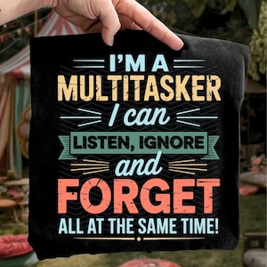Puede incluir: Camiseta negra con el texto "I'M A MULTITASKER I can LISTEN, IGNORE and FORGET ALL AT THE SAME TIME!" en varios colores. El texto está en una fuente en negrita y estilizada. La camiseta está sujeta por una mano.