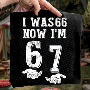 May include: Black t-shirt with the text "I WAS 66 NOW I'M 67" in white, distressed font. The number 67 is large and centered, with two hands at the bottom. The shirt is held up by a hand.