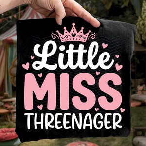 May include: Black t-shirt with the text "Little Miss Threenager" in white and pink. The design includes a pink crown and small pink hearts. The shirt is held up by a hand.