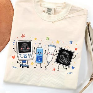 Op de afbeelding: Een crèmekleurig t-shirt met een afbeelding van een lachend echografie-apparaat, een blauwe fles echogel, een transducer en een echoscopiebeeld van een foetus, omringd door kleurrijke, met de hand getekende sterren en hartjes.