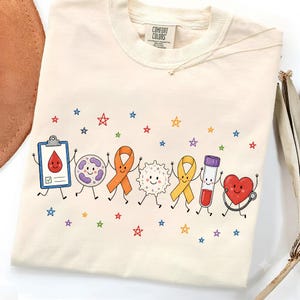 Puede incluir: Una camiseta de color crema con un gráfico de personajes médicos de dibujos animados tomados de la mano, incluyendo un portapapeles con una gota de sangre, una célula, un lazo naranja, una célula blanca, un lazo amarillo y un tubo de ensayo.