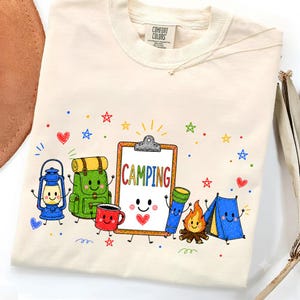 May include: A cream-colored t-shirt featuring a colorful graphic of anthropomorphic camping gear including a lantern, backpack, mug, clipboard with the text CAMPING, flashlight, campfire, and tent, surrounded by small stars and hearts.