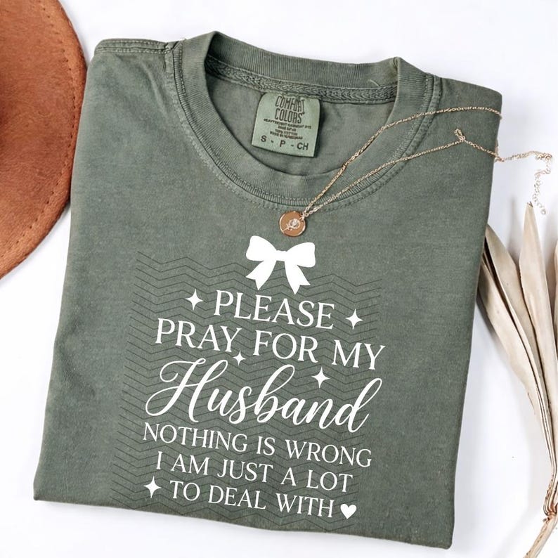 May include: An olive green t-shirt with the text "PLEASE PRAY FOR MY Husband" in white script. The shirt has a small bow graphic and a gold necklace. The shirt is folded and has a Comfort Colors tag.