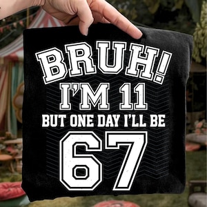 Op de afbeelding: Zwart T-shirt met witte tekst: "BRUH! IK BEN 11 MAAR OP EEN DAG BEN IK 67". De tekst is in een lettertype in collegestijl met een zwarte omtrek. Het T-shirt wordt door een hand omhoog gehouden.