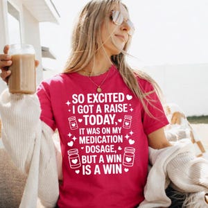 K&ouml;nnte beinhalten: Magentafarbenes T-Shirt mit wei&szlig;em Text: &bdquo;SO EXCITED I GOT A RAISE TODAY, IT WAS ON MY MEDICATION DOSAGE, BUT A WIN IS A WIN&ldquo;. Das Shirt zeigt Herzen und Pillenflaschen-Grafiken.