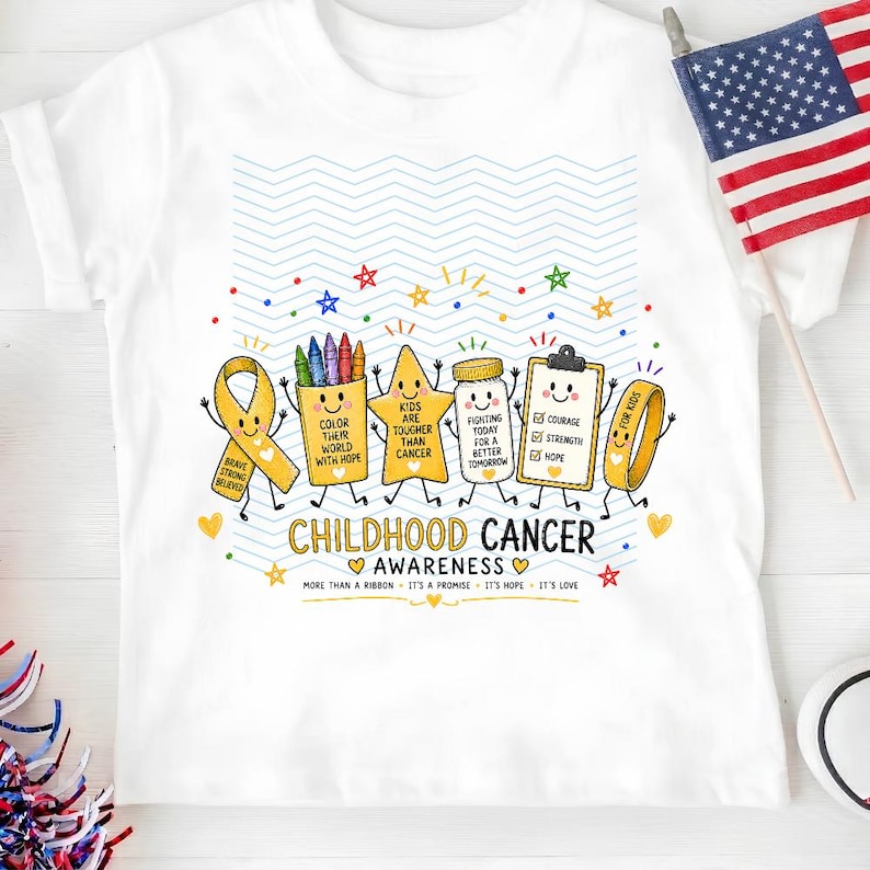 May include: A white t-shirt featuring a colorful graphic of cartoon characters including a yellow ribbon, a crayon box, a star, a medicine bottle, a clipboard, and a wristband. The text reads CHILDHOOD CANCER AWARENESS and includes motivational phrases.