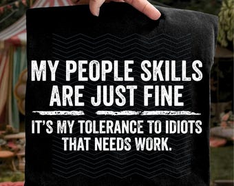 Peut inclure: Coussin noir avec texte blanc : "MY PEOPLE SKILLS ARE JUST FINE. IT'S MY TOLERANCE TO IDIOTS THAT NEEDS WORK." Le coussin est tenu par une main, avec un arrière-plan extérieur flou.