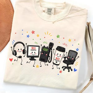May include: A cream-colored t-shirt featuring a graphic of five anthropomorphic 911 dispatcher items: a headset, a computer monitor labeled 911 DISPATCH, a radio, a coffee cup labeled 911 DISPATCH, and an office chair, surrounded by colorful stars.