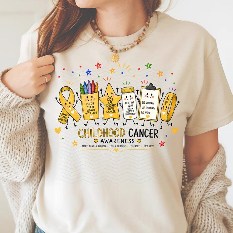 May include: A beige t-shirt featuring a graphic of childhood cancer awareness symbols with faces, including a yellow ribbon, crayons, a star, a medicine bottle, and a clipboard. Text reads: CHILDHOOD CANCER AWARENESS, MORE THAN A RIBBON.