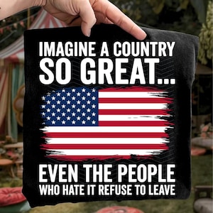 May include: Black t-shirt featuring the American flag with a distressed design. The text reads "IMAGINE A COUNTRY SO GREAT... EVEN THE PEOPLE WHO HATE IT REFUSE TO LEAVE" in white lettering. The shirt is held up by a hand.
