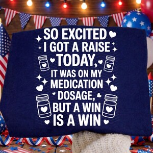 K&ouml;nnte beinhalten: Marineblaues T-Shirt mit wei&szlig;em Text: "SO EXCITED I GOT A RAISE TODAY, IT WAS ON MY MEDICATION DOSAGE, BUT A WIN IS A WIN." Das Design umfasst Herzen, Sterne und Pillenflaschen-Grafiken. Patriotisches Thema.