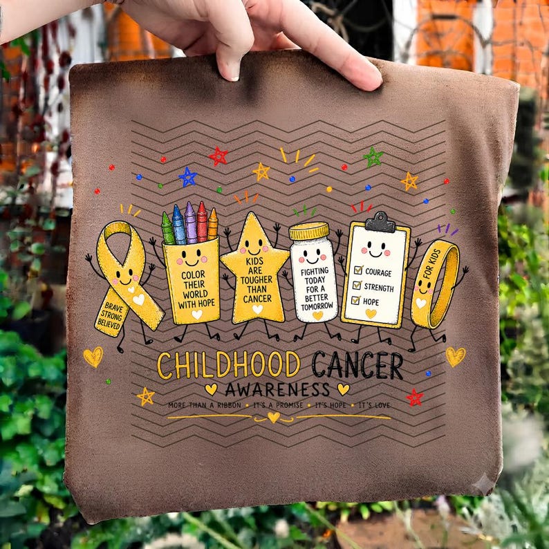 May include: A brown t-shirt featuring a Childhood Cancer Awareness graphic with a yellow ribbon, a crayon cup, a star, a medicine jar, a clipboard, and a wristband. The text reads: Color their world with hope, Kids are tougher than cancer, Fighting today for a better tomorrow, Courage, Strength, Hope, For kids, and More than a ribbon.