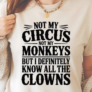 Może przedstawiać: Beżowa koszulka z czarnym napisem: "NOT MY CIRCUS NOT MY MONKEYS BUT I DEFINITELY KNOW ALL THE CLOWNS". Top ma okrągły dekolt i krótkie rękawy. Widoczny jest naszyjnik z zawieszką w kształcie słońca.