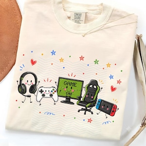 May include: A cream-colored t-shirt featuring a graphic print of anthropomorphic gaming equipment including a headset, a game controller, a computer monitor with the text GAME ON, a gaming chair, and a handheld console, surrounded by colorful stars.