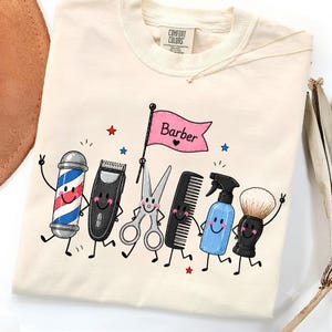 以下が含まれることがあります： クリーム色のTシャツで、バーバーポール、バリカン、ハサミ、コーム、スプレーボトル、シェービングブラシといった擬人化された理容道具のグラフィックが描かれています。道具の上にあるピンクの旗には、ハートマークと共に「Barber」という文字が記されています。