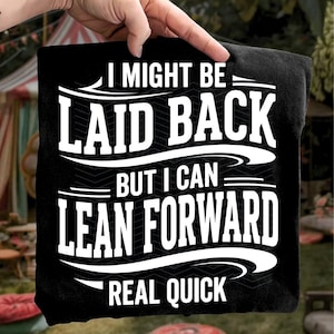 May include: Black t-shirt with white text that reads "I MIGHT BE LAID BACK BUT I CAN LEAN FORWARD REAL QUICK". The shirt is held up by a hand, with a blurred outdoor background.
