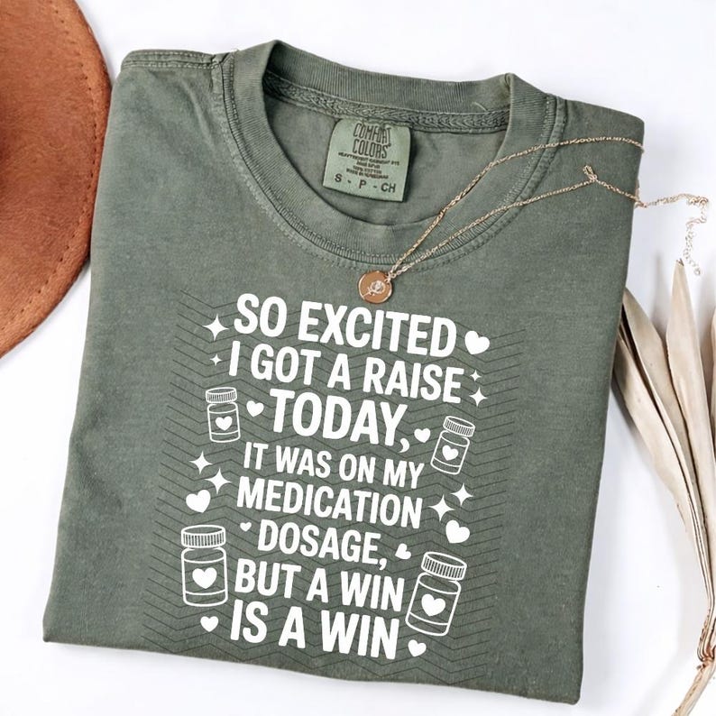 K&ouml;nnte beinhalten: Olivgr&uuml;nes T-Shirt mit dem wei&szlig;en Text "SO EXCITED I GOT A RAISE TODAY, IT WAS ON MY MEDICATION DOSAGE, BUT A WIN IS A WIN". Das T-Shirt hat einen Rundhalsausschnitt und ein kleines Etikett. Eine goldene Halskette liegt auf dem T-Shirt.