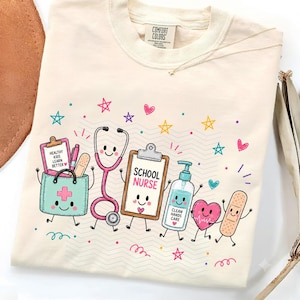 May include: A cream-colored t-shirt featuring a colorful graphic of a school nurse theme. The design includes a medical bag, a stethoscope, a clipboard labeled 'SCHOOL NURSE', a hand sanitizer bottle labeled 'CLEAN HANDS CARE', a heart, and a bandage.
