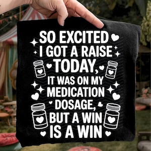 K&ouml;nnte beinhalten: Schwarzes T-Shirt mit wei&szlig;em Text: "SO EXCITED I GOT A RAISE TODAY, IT WAS ON MY MEDICATION DOSAGE, BUT A WIN IS A WIN." Das Design umfasst Pillenflaschen-Grafiken mit Herzen, Sternen und Herzen.