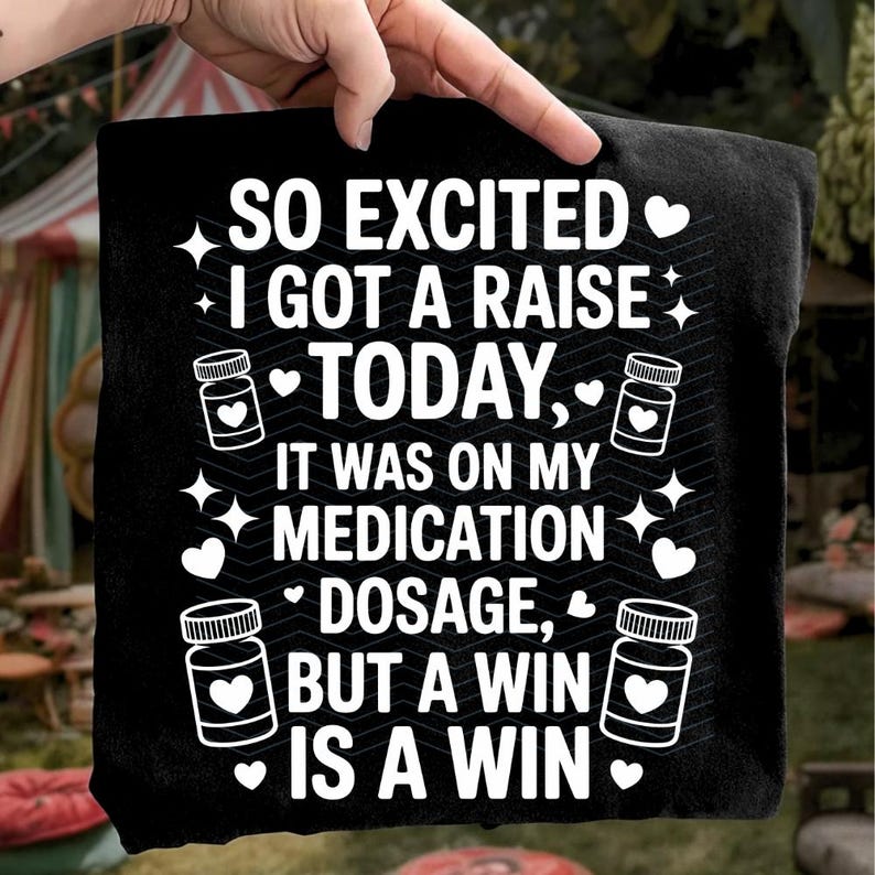 K&ouml;nnte beinhalten: Schwarzes T-Shirt mit wei&szlig;em Text: "SO EXCITED I GOT A RAISE TODAY, IT WAS ON MY MEDICATION DOSAGE, BUT A WIN IS A WIN." Das Design umfasst Pillenflaschen-Grafiken mit Herzen, Sternen und Herzen.
