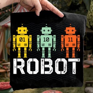May include: Black t-shirt featuring three colorful robot illustrations. The robots are yellow, mint green, and orange, each with a unique number. The word "ROBOT" is printed in large white letters below the robots.