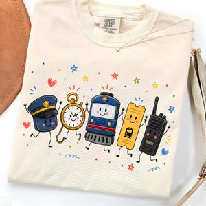 Könnte beinhalten: Ein cremefarbenes T-Shirt mit einem Grafikdruck von fünf anthropomorphen Charakteren: eine blaue Schaffnermütze, eine goldene Taschenuhr, eine blaue Zuglokomotive, ein gelbes Zugticket und ein schwarzes Funkgerät, umgeben von bunten Sternen und Herzen.