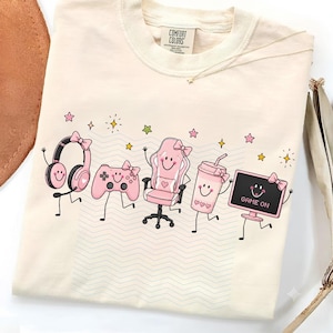 Può includere: Una t-shirt color crema con una stampa grafica di cinque oggetti da gioco antropomorfi in rosa, tra cui cuffie, un controller, una sedia, un bicchiere e un monitor con il testo GAME ON, il tutto accentuato da piccole stelle.