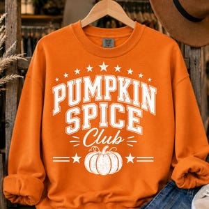 May include: An orange long-sleeve sweatshirt featuring white screen-printed text that reads PUMPKIN SPICE Club in a collegiate font style. The design includes a graphic of a pumpkin centered below the text, framed by stars and horizontal lines.