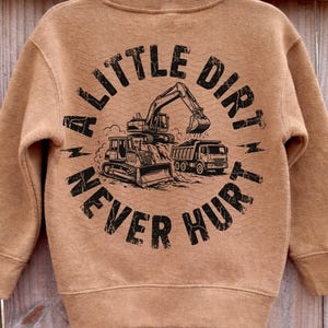 May include: A brown sweatshirt with the text "A Little Dirt Never Hurt" in a circular design. The design includes illustrations of construction vehicles such as an excavator, bulldozer, and dump truck.
