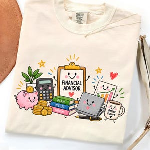 May include: A cream-colored t-shirt featuring a colorful graphic of a smiling clipboard labeled "FINANCIAL ADVISOR" surrounded by a pink piggy bank, gold coins, a calculator, a stack of books labeled "PLAN," "INVEST," "GROW," a laptop, a chart, and a mug.