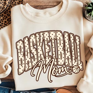 Könnte beinhalten: Ein cremefarbenes Sweatshirt mit Rundhalsausschnitt, das das Wort BASKETBALL in großen, braunen, gepunkteten Blockbuchstaben zeigt. Darunter steht das Wort Mom in brauner Schreibschrift, wobei ein kleiner Basketball das o ersetzt.