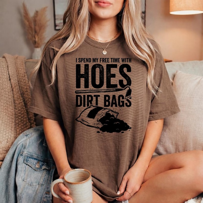 May include: A brown t-shirt featuring the text "I spend my free time with HOES DIRT BAGS" in black. The graphic includes a bag of dirt and gardening tools. The shirt is a casual style.