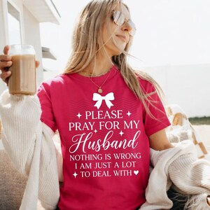 May include: A magenta t-shirt with the text "PLEASE PRAY FOR MY Husband" in white script. The shirt also includes the phrases "NOTHING IS WRONG I AM JUST A LOT TO DEAL WITH". The shirt has a bow graphic.