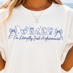May include: A white t-shirt featuring a blue graphic print of pharmacy-themed icons including bows, a mortar and pestle, a pill bottle, capsules, a clipboard, and a caduceus, with the text "I'm Literally Just A pharmacist" written in blue script.