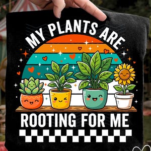 Op de afbeelding: Zwart t-shirt met een kleurrijke afbeelding van potplanten en de tekst "MY PLANTS ARE ROOTING FOR ME". Het ontwerp toont een vetplant, twee bladplanten en een zonnebloem, allemaal met lachende gezichten.