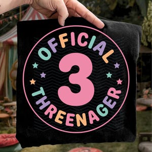 May include: Black t-shirt with a colorful circular design. The design features the number "3" in pink, surrounded by the words "OFFICIAL THREENAGER" in various colors, with star accents. The shirt is held up by a hand.