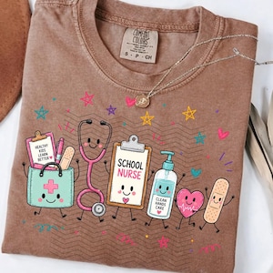 May include: A brown t-shirt featuring a graphic of a medical bag, stethoscope, clipboard labeled SCHOOL NURSE, hand sanitizer, heart, and bandage. The medical bag has a clipboard with the text HEALTHY KIDS LEARN BETTER. Colorful stars surround the items.