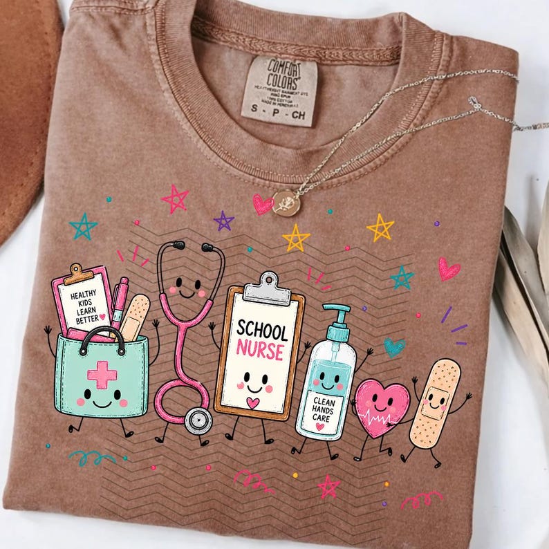May include: A brown t-shirt featuring a graphic of a medical bag, stethoscope, clipboard labeled SCHOOL NURSE, hand sanitizer, heart, and bandage. The medical bag has a clipboard with the text HEALTHY KIDS LEARN BETTER. Colorful stars surround the items.