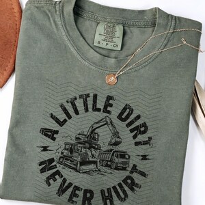 May include: Olive green t-shirt with the text "A Little Dirt Never Hurt" and construction vehicle graphics. A gold necklace with a pendant rests on the shirt. The shirt is a size small.