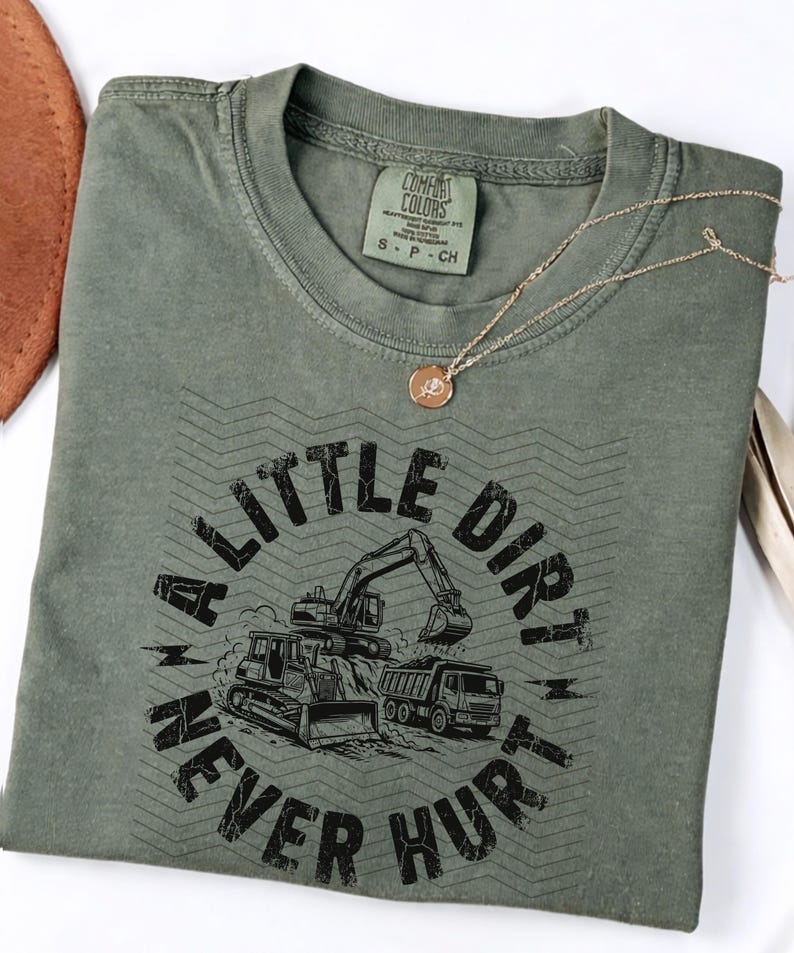 May include: Olive green t-shirt with the text "A Little Dirt Never Hurt" and construction vehicle graphics. A gold necklace with a pendant rests on the shirt. The shirt is a size small.