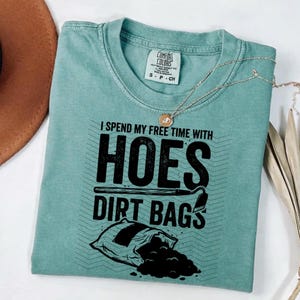 May include: A teal t-shirt with the text "I spend my free time with hoes dirt bags" in black. The shirt features a graphic of a shovel and a bag of dirt. A silver necklace with a pendant is draped over the shirt.