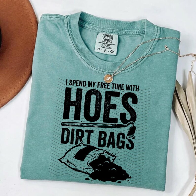 May include: A teal t-shirt with the text "I spend my free time with hoes dirt bags" in black. The shirt features a graphic of a shovel and a bag of dirt. A silver necklace with a pendant is draped over the shirt.