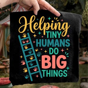 May include: Black t-shirt with the text "Helping Tiny Humans Do Big Things" in yellow, teal, and coral. A teal ladder with handprints is on the left side of the design. The shirt is held up by a hand.