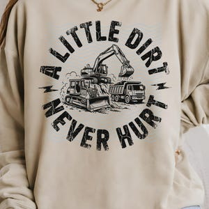 May include: Beige sweatshirt with a black and white graphic featuring construction vehicles: a bulldozer, excavator, and dump truck. The text "A LITTLE DIRT NEVER HURT" is printed in a distressed font, encircling the vehicles.