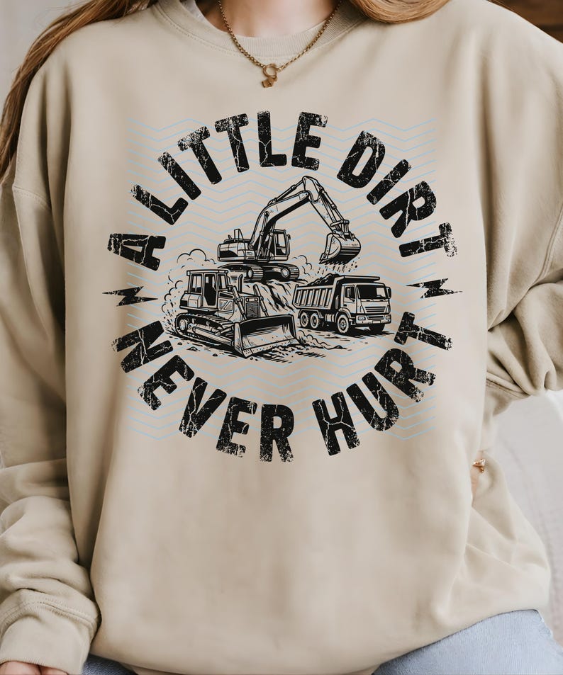 May include: Beige sweatshirt with a black and white graphic featuring construction vehicles: a bulldozer, excavator, and dump truck. The text "A LITTLE DIRT NEVER HURT" is printed in a distressed font, encircling the vehicles.
