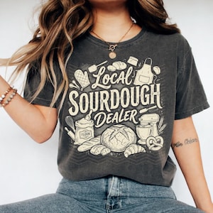 May include: Dark grey t-shirt with a cream-colored graphic that reads "Local Sourdough Dealer." The design includes illustrations of bread, a mixer, flour, and baking tools. The shirt is a casual, comfortable style.