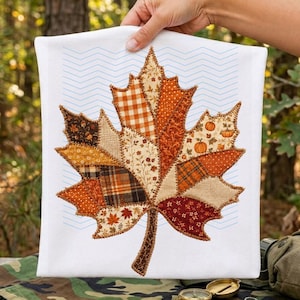 May include: A white fabric towel featuring a large, central maple leaf applique made from various patchwork fabric scraps in shades of orange, brown, and cream. The leaf is outlined with dark brown stitching and set against a light blue chevron pattern.