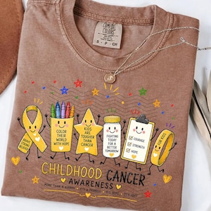 May include: A brown t-shirt featuring a graphic with a yellow awareness ribbon, a crayon cup, a star, a medicine bottle, and a clipboard. The text reads "Childhood Cancer Awareness" along with "Color their world with hope," "Kids are tougher than cancer," and "Fighting today for a better tomorrow."