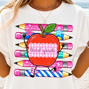 Könnte beinhalten: Ein weißes T-Shirt mit einer Grafik eines roten Apfels mit grünem Blatt und dünnen schwarzen Gliedmaßen. Der Apfel hat ein rosa-weiß kariertes Etikett mit dem Text third grade. Dahinter befinden sich horizontal angeordnete rosa und blaue Stifte.