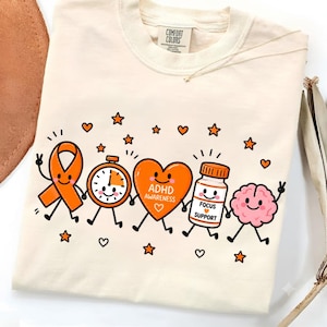 Pode incluir: Uma t-shirt cor de creme com um gráfico de cinco personagens de desenhos animados de mãos dadas: uma fita de consciencialização laranja, um relógio, um coração laranja com o texto ADHD AWARENESS, um frasco de comprimidos e um cérebro rosa.